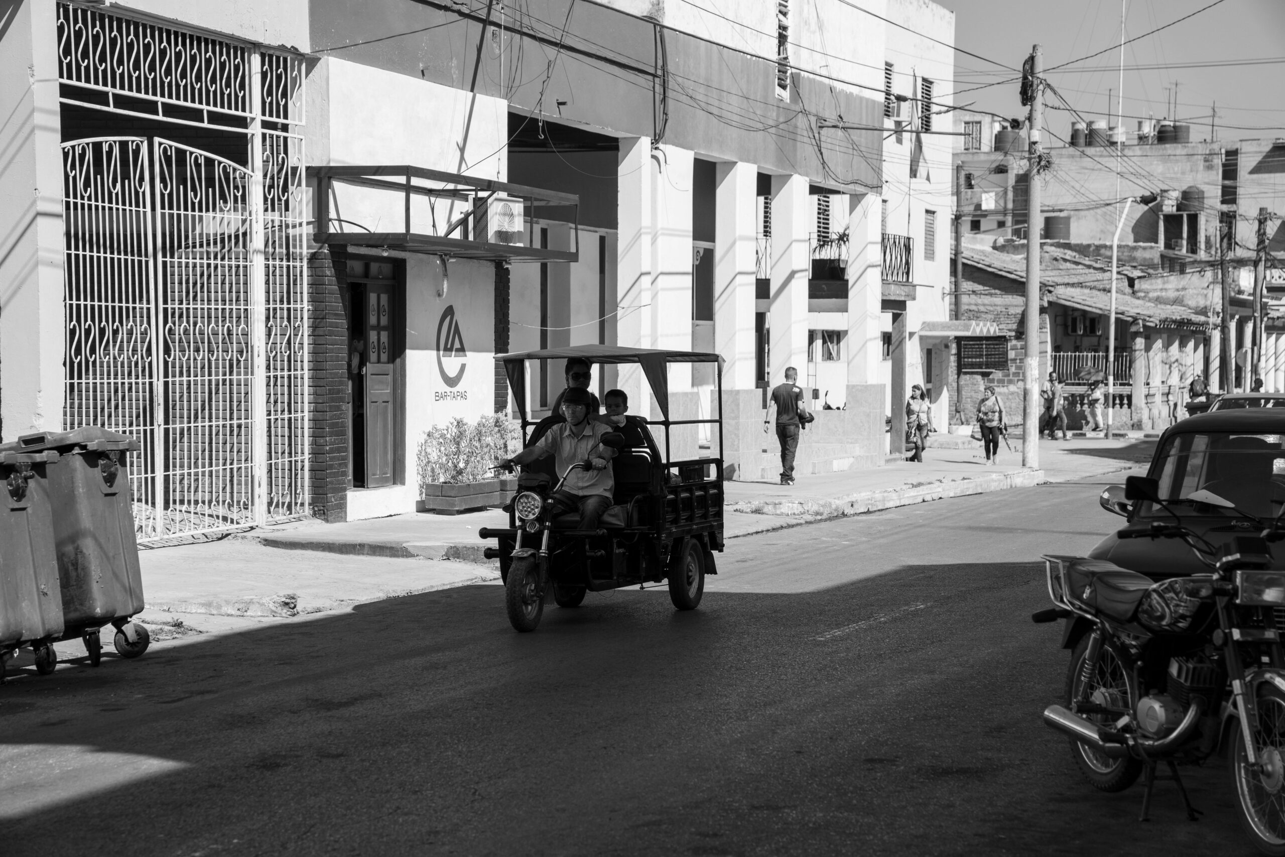 Black and white street view in Pinar del Río, featuring a rikshaw and pedestrians on a sunny day.